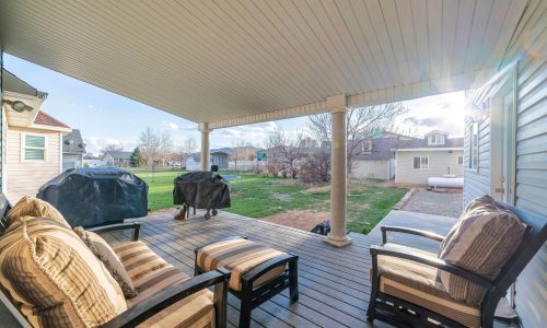 Covered patio of a house with wooden couch and two covered grills. There is a wooden planks flooring and a view of a lawn and neighborhood houses.